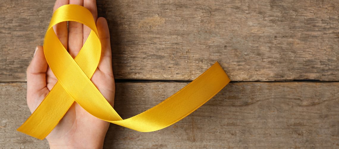 Yellow awareness ribbon in female hand on wooden background